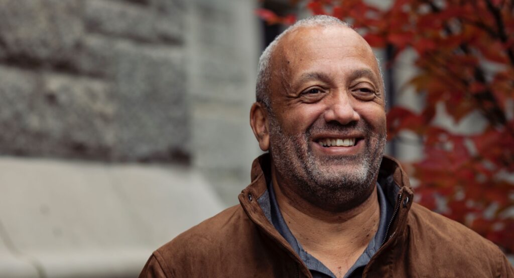 John Swinton is a middle-aged black man wearing a blazer and smiling