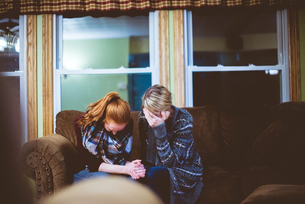 a young adult female and middle-aged adult female sit on a couch holding hands, their heads bent in grief.