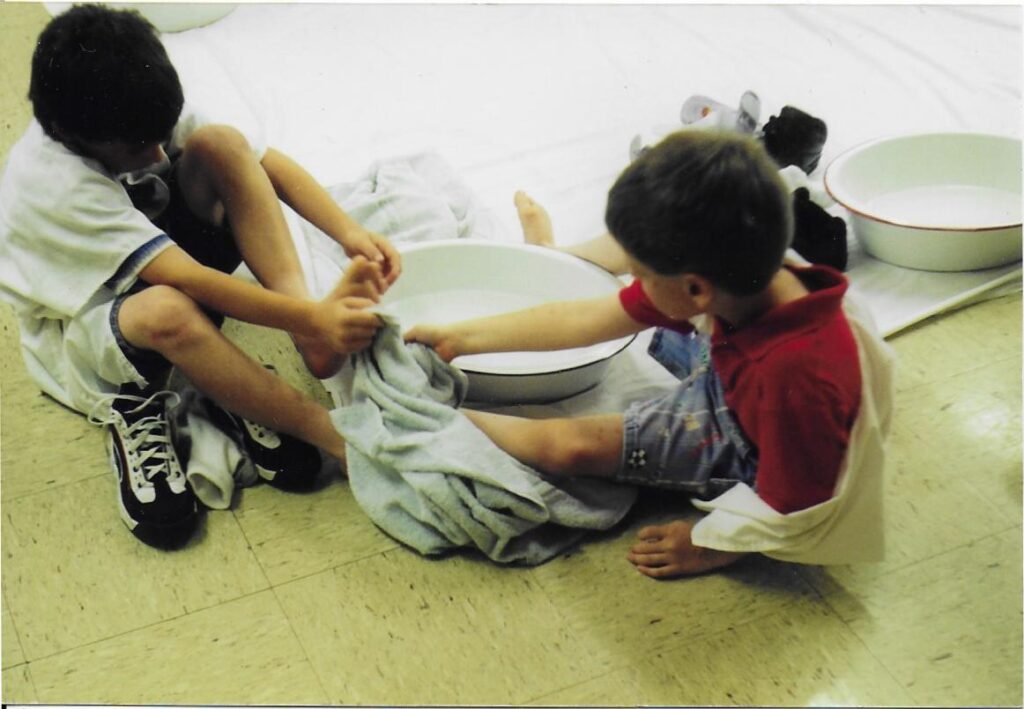 a young boy washes another boy's feet using a basin and towel. In the background are one boy's leg braces.