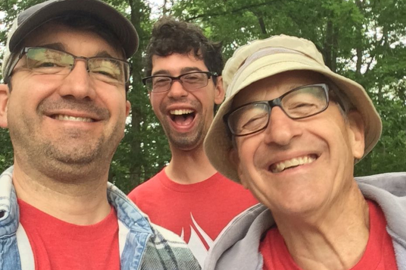 Three white men smile joyfully: one is a young adult with a visible sign of a disability, one is a middle-aged man, and one is an older male.