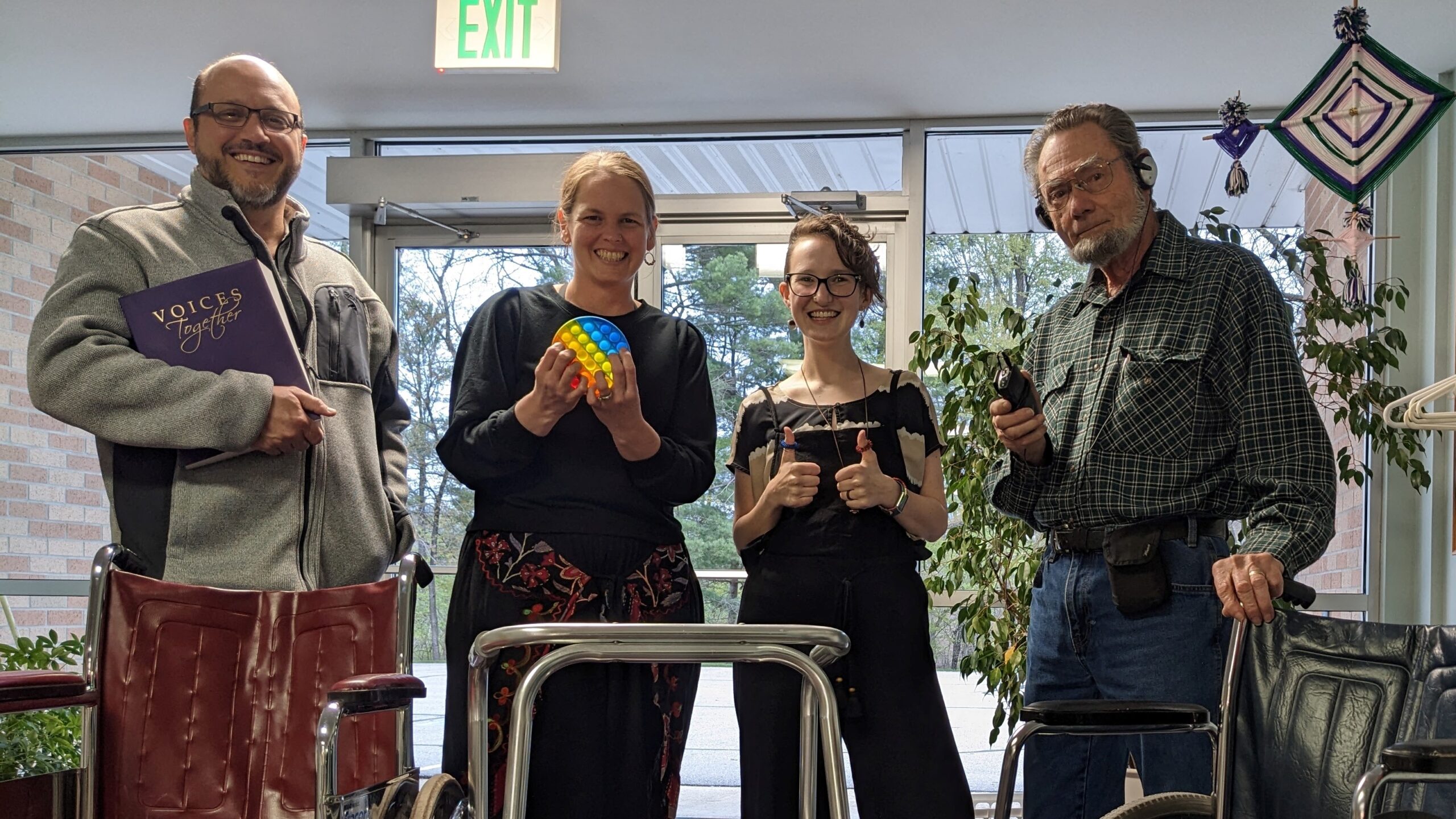 Four white adults, ranging from young adult to older adult, hold accessibility tools and stand behind wheelchairs.