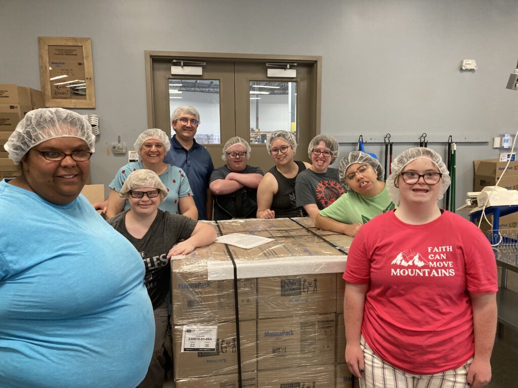 seven young adults of various ethnicities and abilities stand around a large cardboard box.