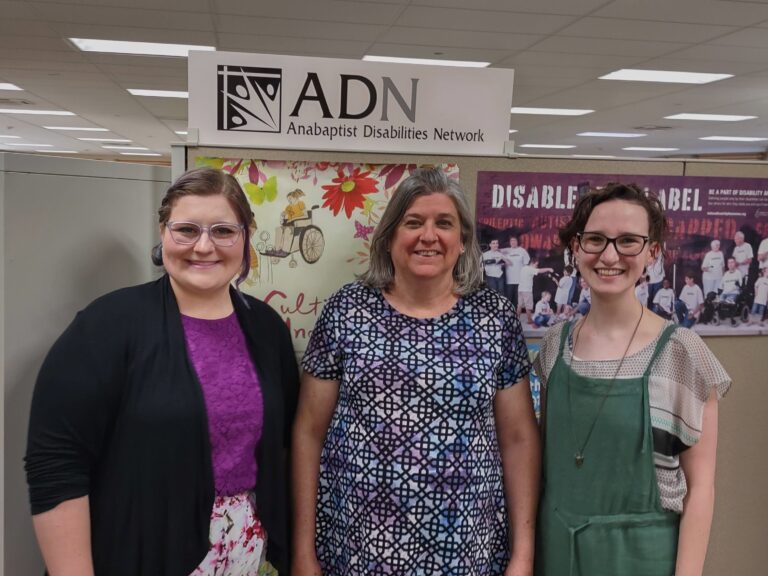 Three white females stand in front of a sign reading "ADN." Zoe and Emily are young females wearing glasses; Jeanne is a middle-aged female with graying hair.