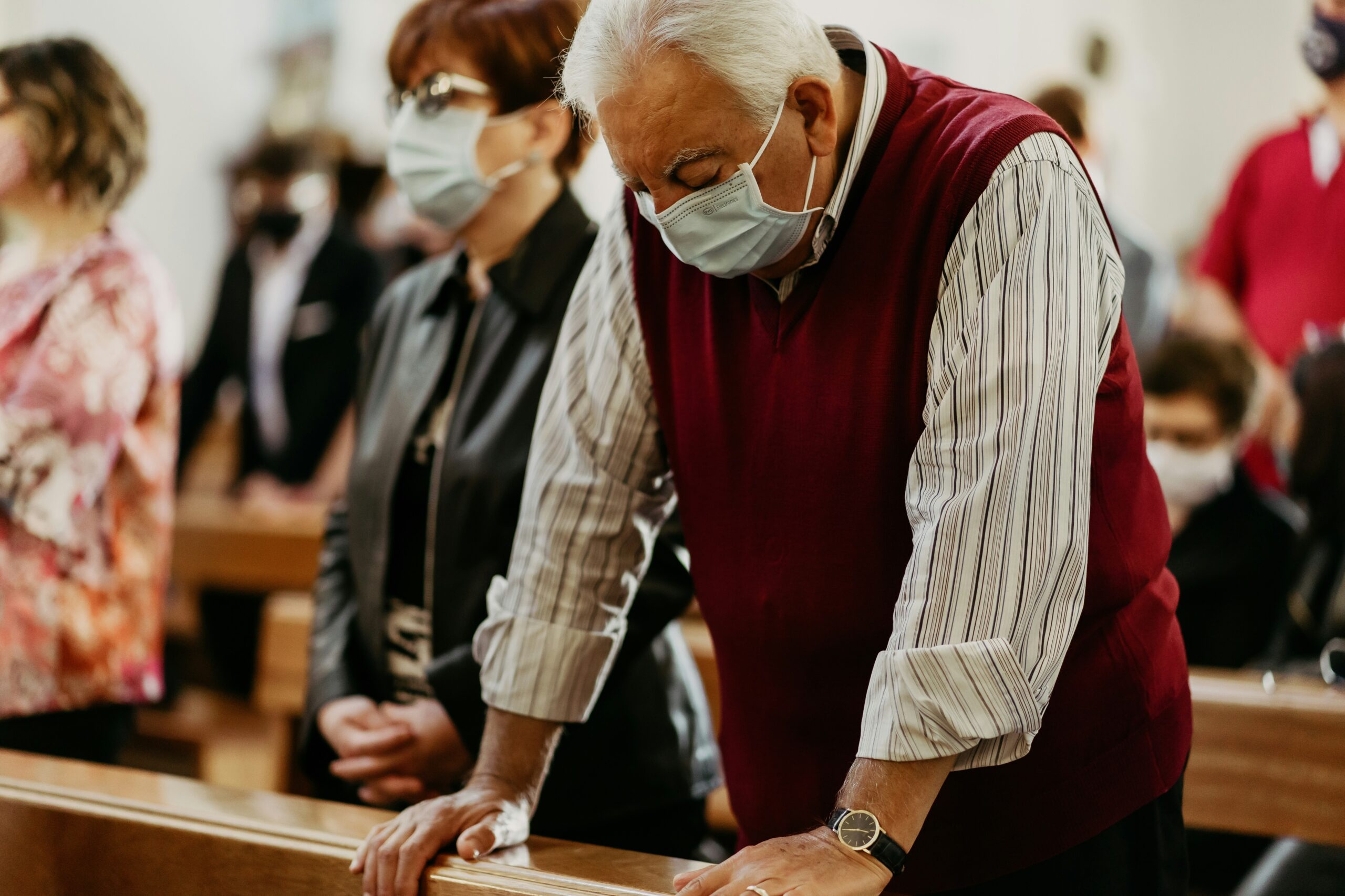 a man and woman stand in church pews. both wear surgical masks.