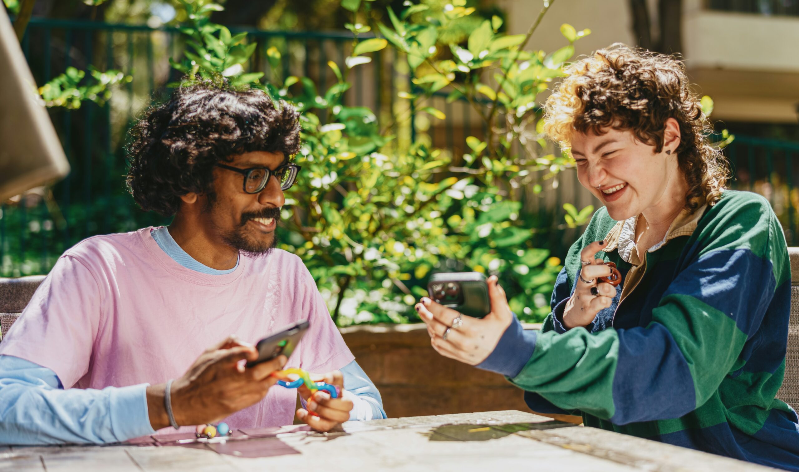 two adults sit outside. One person shows their phone to the other person. Both people are smiling.