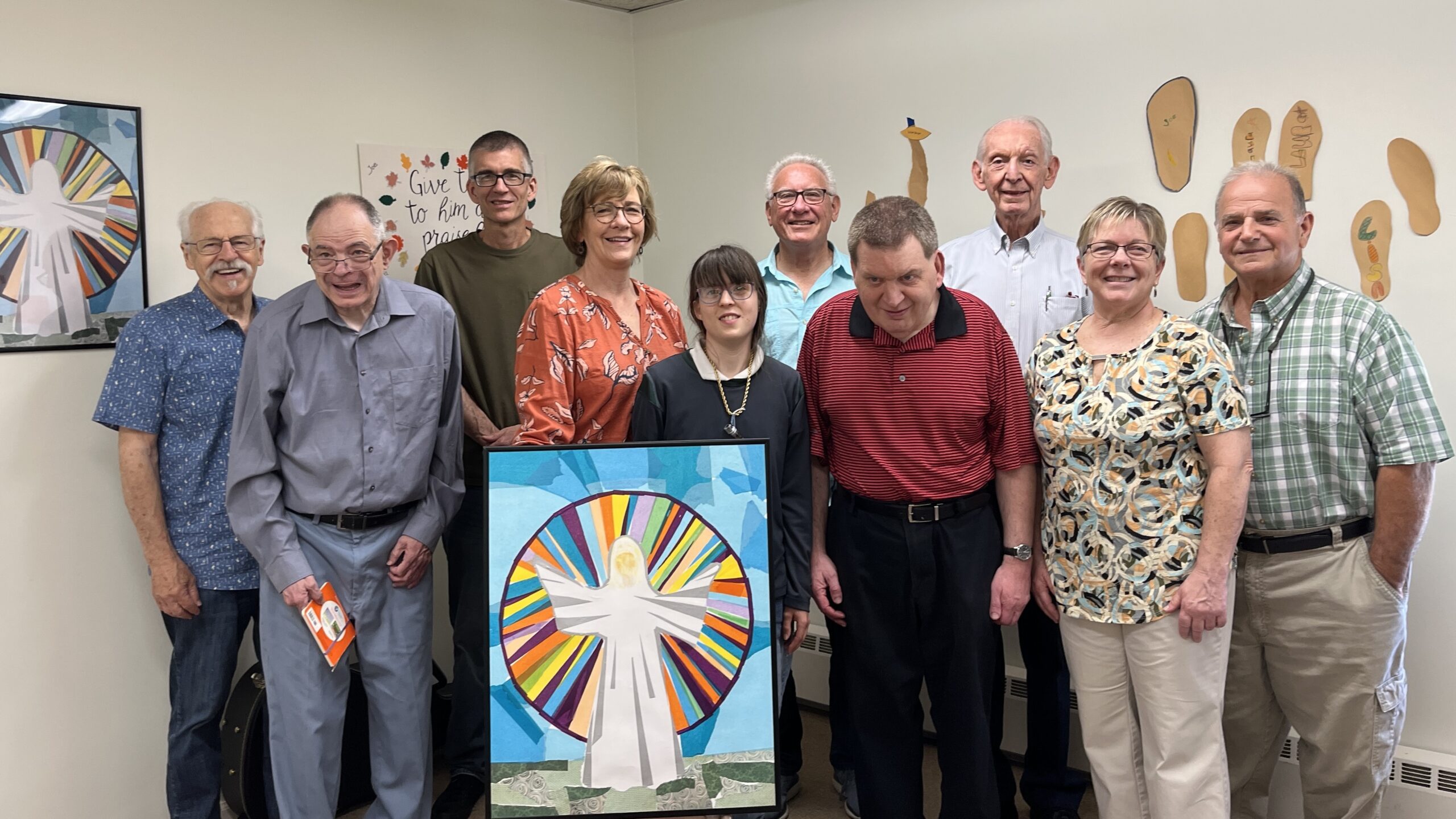 a group of white adults of various genders. Some have visible signs of intellectual disabilities. A person in the middle is holding a large depiction of the Transfiguration made of brightly colored torn papers.