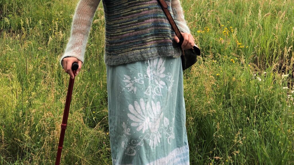 a person in a knit sweater and long skirt lean on a cane in the middle of a field.