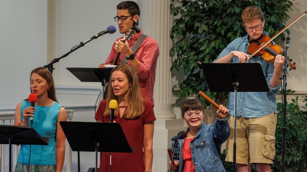 Four white young adults and one white youth play instruments and sing on a stage. The youth has visible signs of down syndrome.