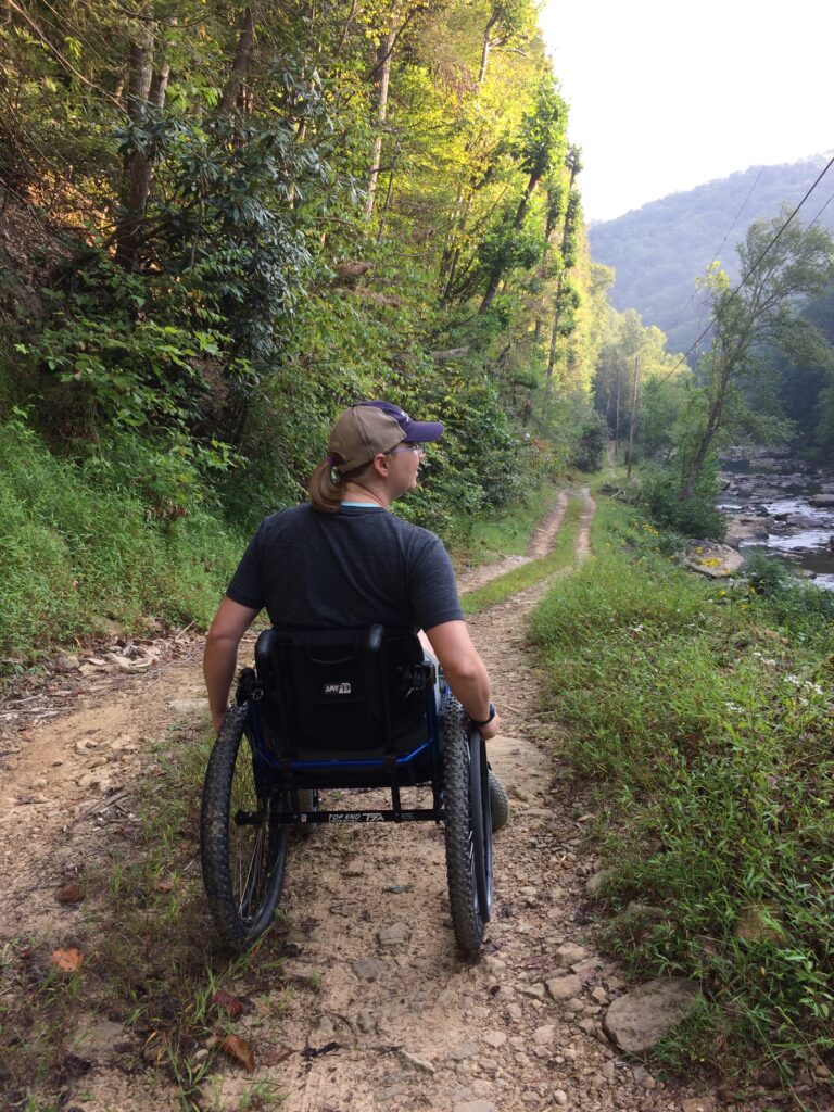 a woman in a wheelchair on a dirt path that cuts through a forest.