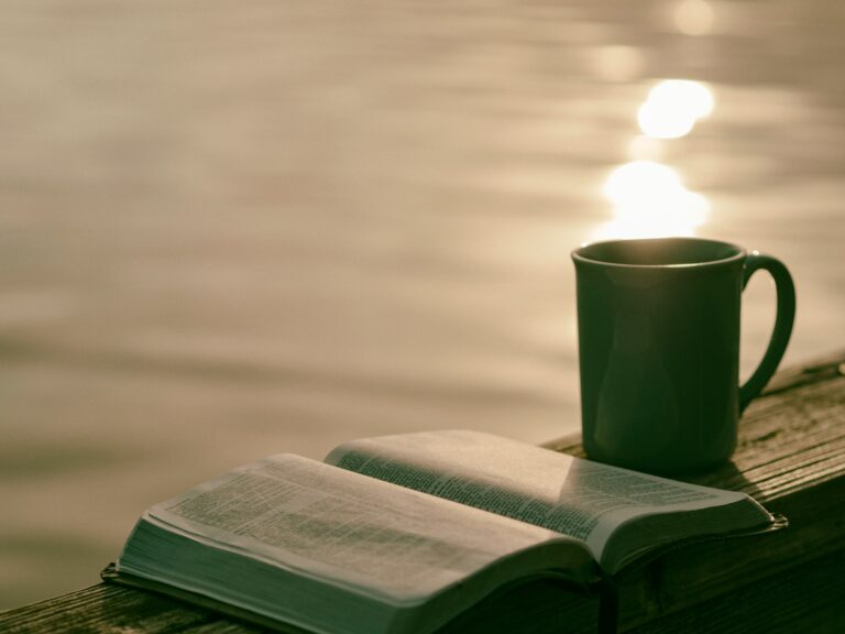 an open bible and a mug of coffee. In the background is a sunrise over a lake.