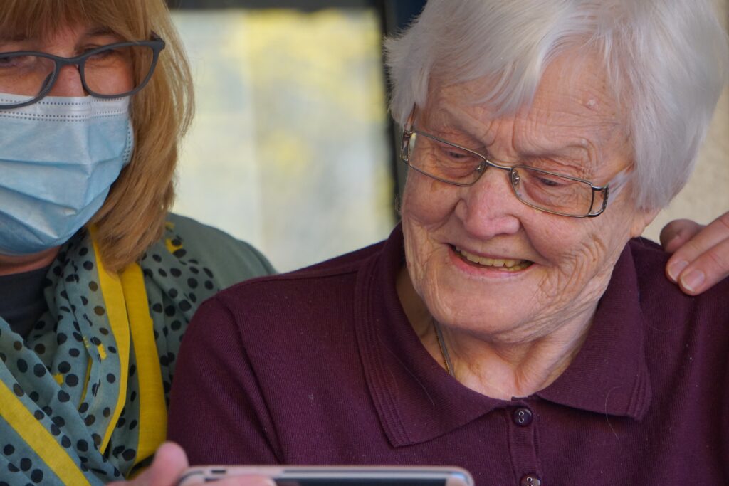 an elderly white woman looks at a phone with a middle-aged woman wearing a mask.