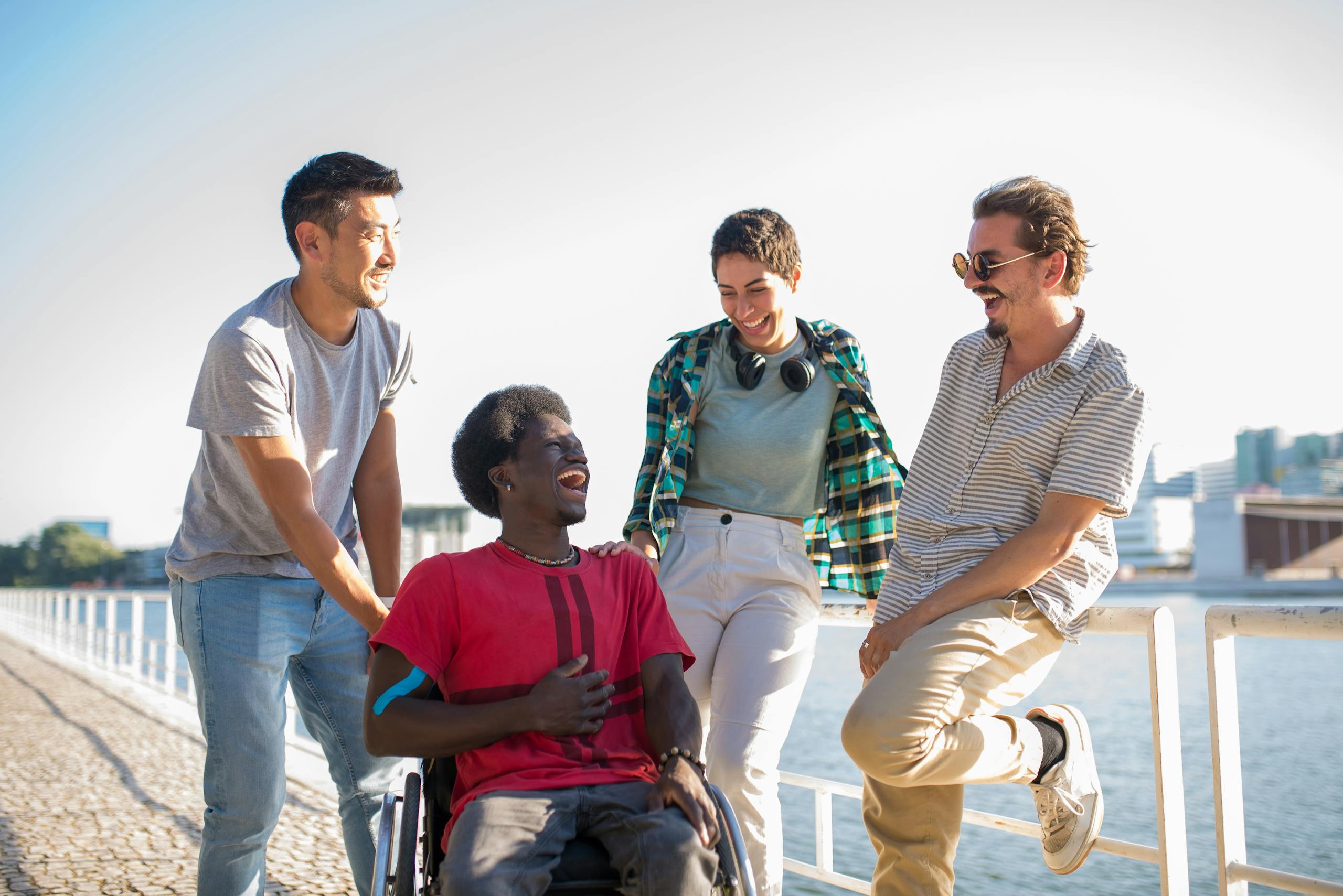 four young adults of various ethnicities talk and laugh together. One person is in a wheelchair and another is wearing noise-reducing headphones