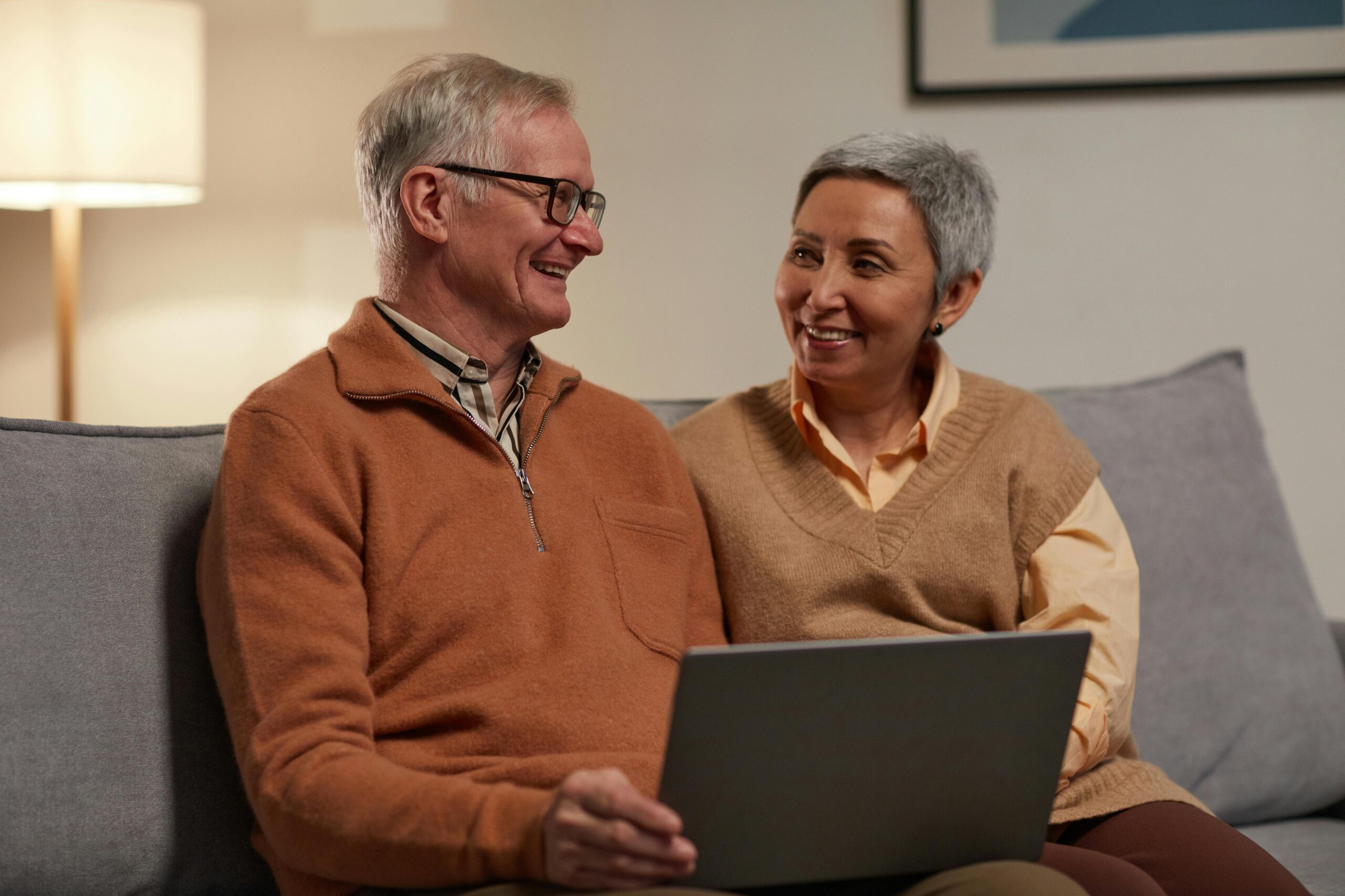 two elderly people sit on a couch with a laptop on their lap, smiling.