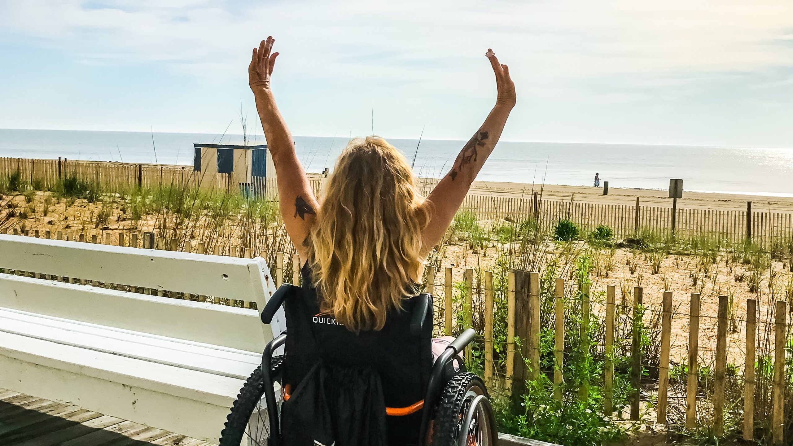 a woman in a wheelchair raises her hands to the sky. In the background is the beach.