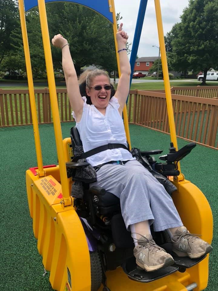 a middle-aged woman in a wheelchair on an accessible swing raises her arms with a look of joy on her face