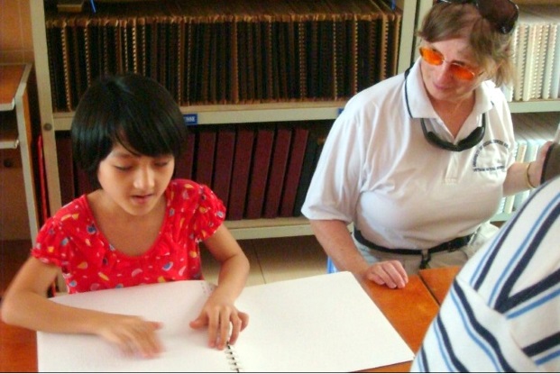 girl with short dark hair and bangs is reading a large Braille book set on the table before her. A woman seated next to her, wearing wire-rimmed glasses with orange lenses, is looking at the girl and smiling