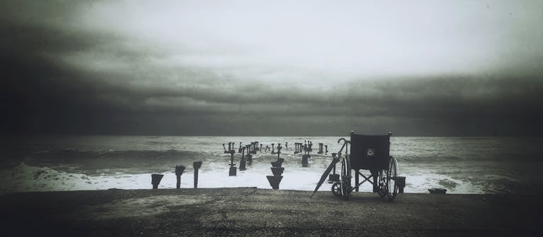 Monochrome image of a lonely wheelchair facing the vast ocean under a dreary sky.